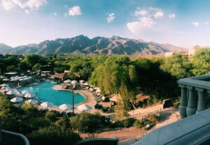 Aerial view of a resort pool surrounded by trees, with mountains in the background under a sunny sky.