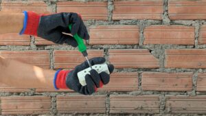 Electrician's gloved hands wiring an electrical outlet with a screwdriver.