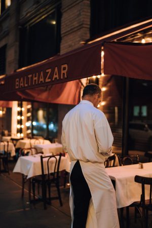 Restaurant storefront at night with a server at the entrance and diners on the patio